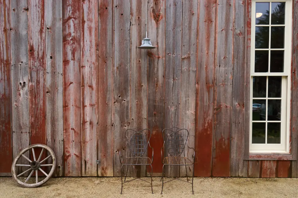 Weathered vertical barn siding in rust and grey, with an old wooden wagon wheel, a small lantern and two empty metal chairs standing against the wall